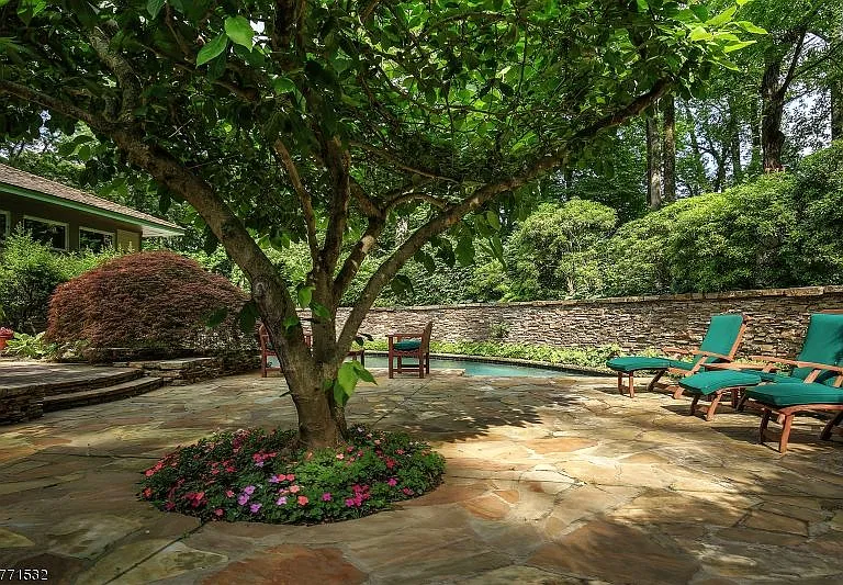 Flagstone patio with a shade tree, green-cushioned lounge chairs, and a pool beside a low stone wall.