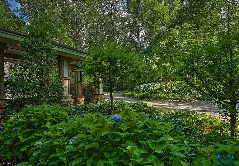 Garden beds of hydrangeas and hostas beside the house's covered walkway, with a gravel drive and tall trees.