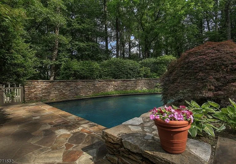 In-ground pool surrounded by a flagstone patio and stone wall, backed by tall trees and a red Japanese maple.